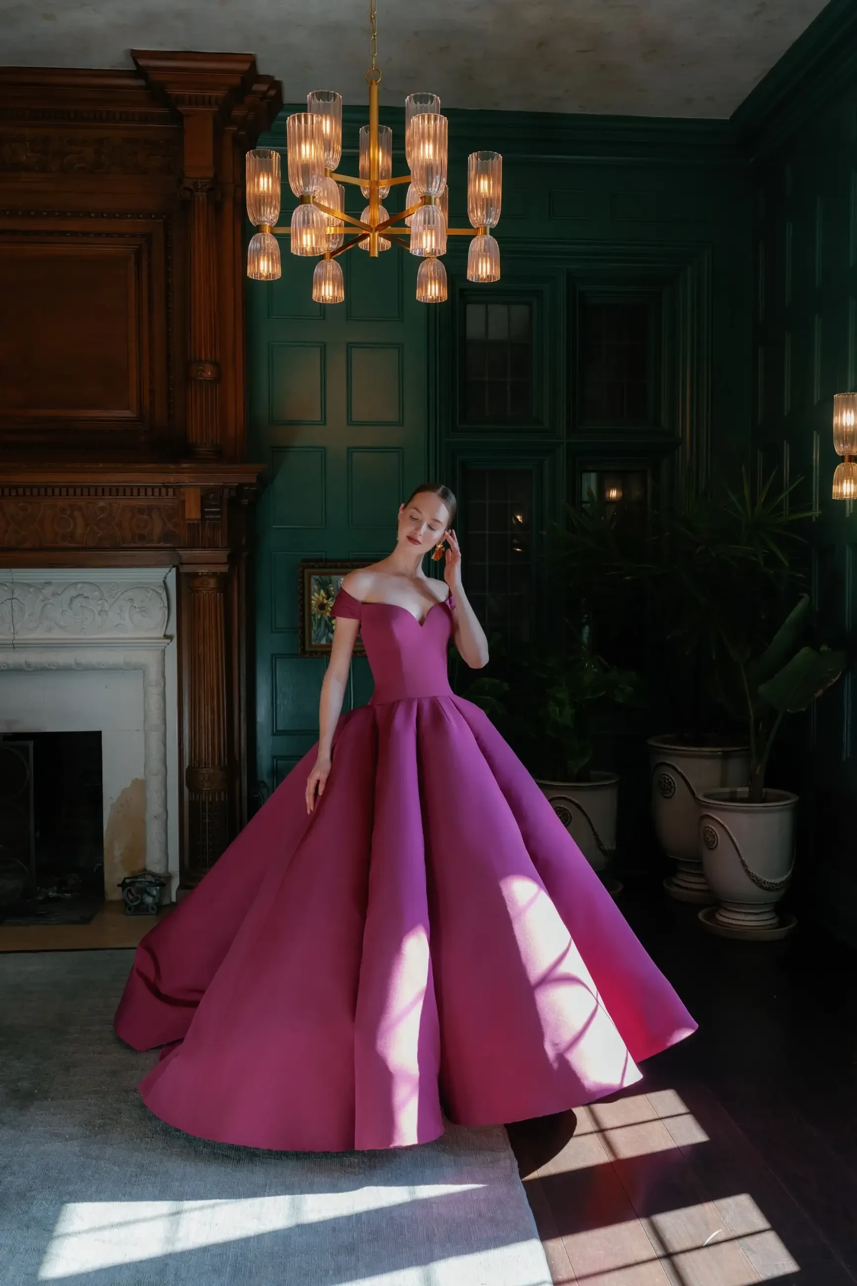A woman in a stunning, off-the-shoulder, magenta ball gown stands in an elegant room with dark green walls and a chandelier, holding a phone to her ear.