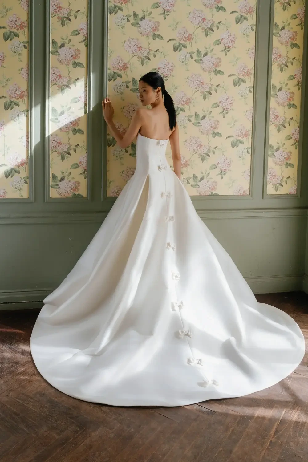 A woman in a strapless white wedding gown, standing with her back to the camera, showcasing a long train and button details. The background features floral-patterned wallpaper and soft lighting.