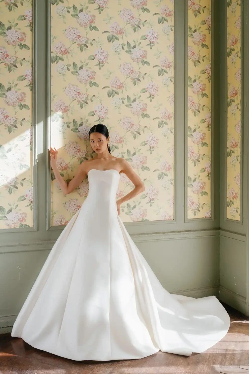 A model wearing a elegant white dress stands against a floral wallpaper backdrop.