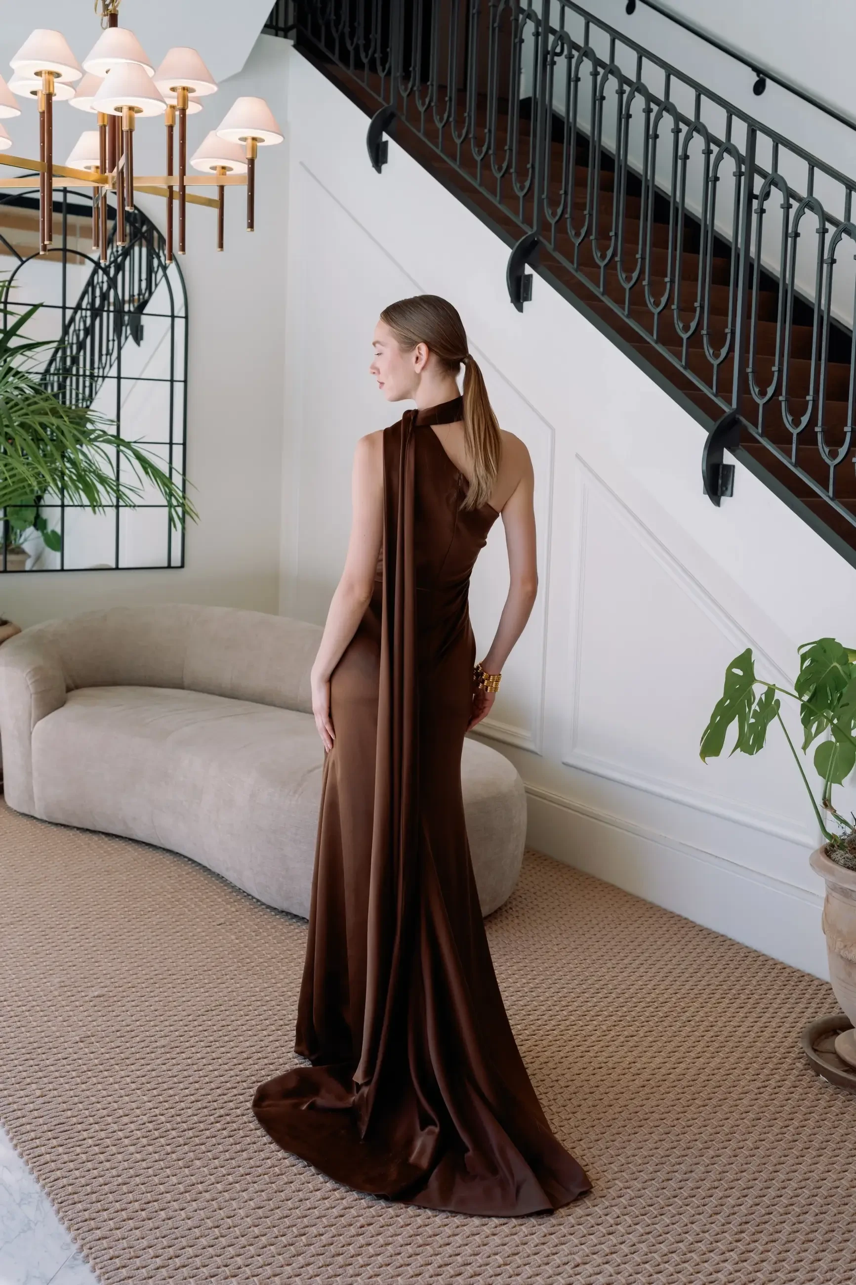 A woman in a brown gown standing with her back to the camera, in a modern interior featuring stairs, a couch, and plants.