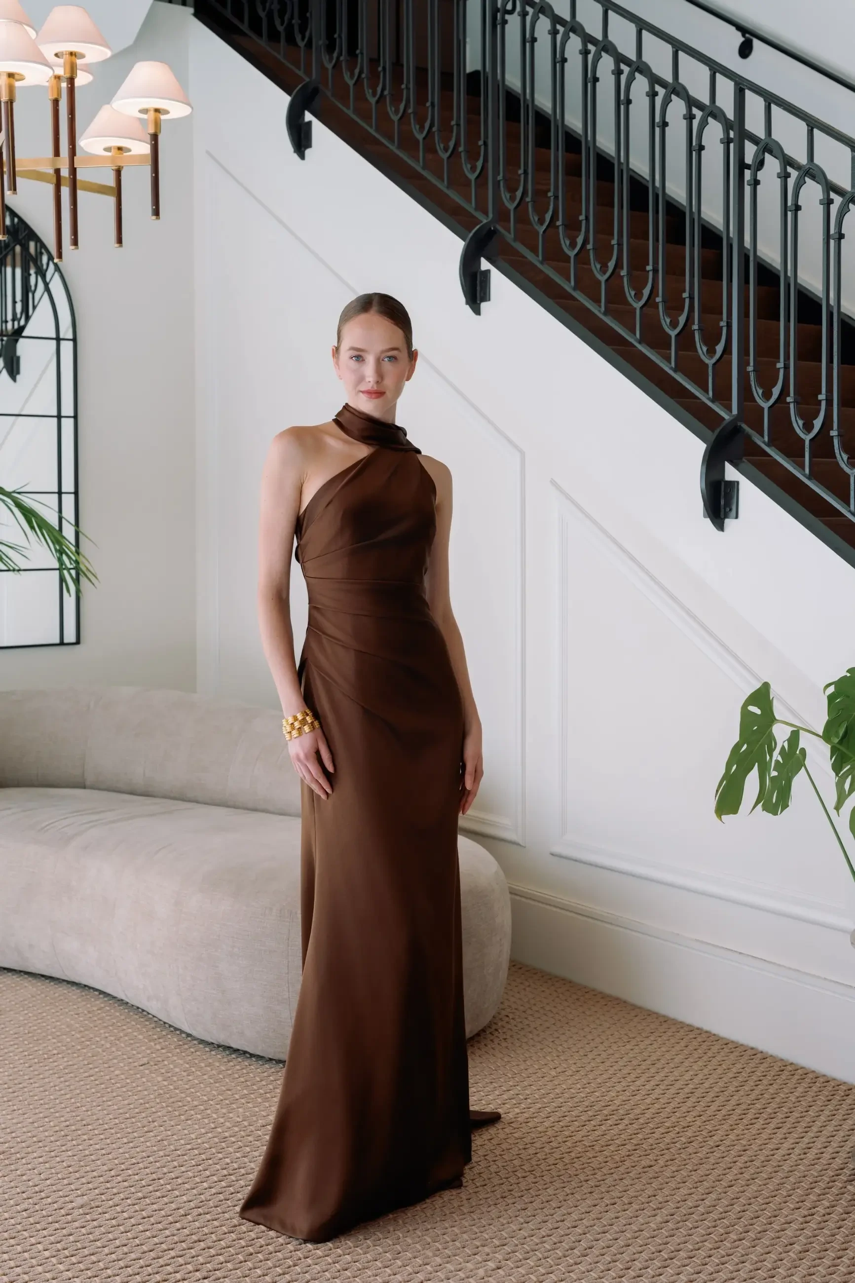 A model wearing a brown evening gown stands on a carpeted floor in an elegant interior space with a staircase and decorative lighting.
