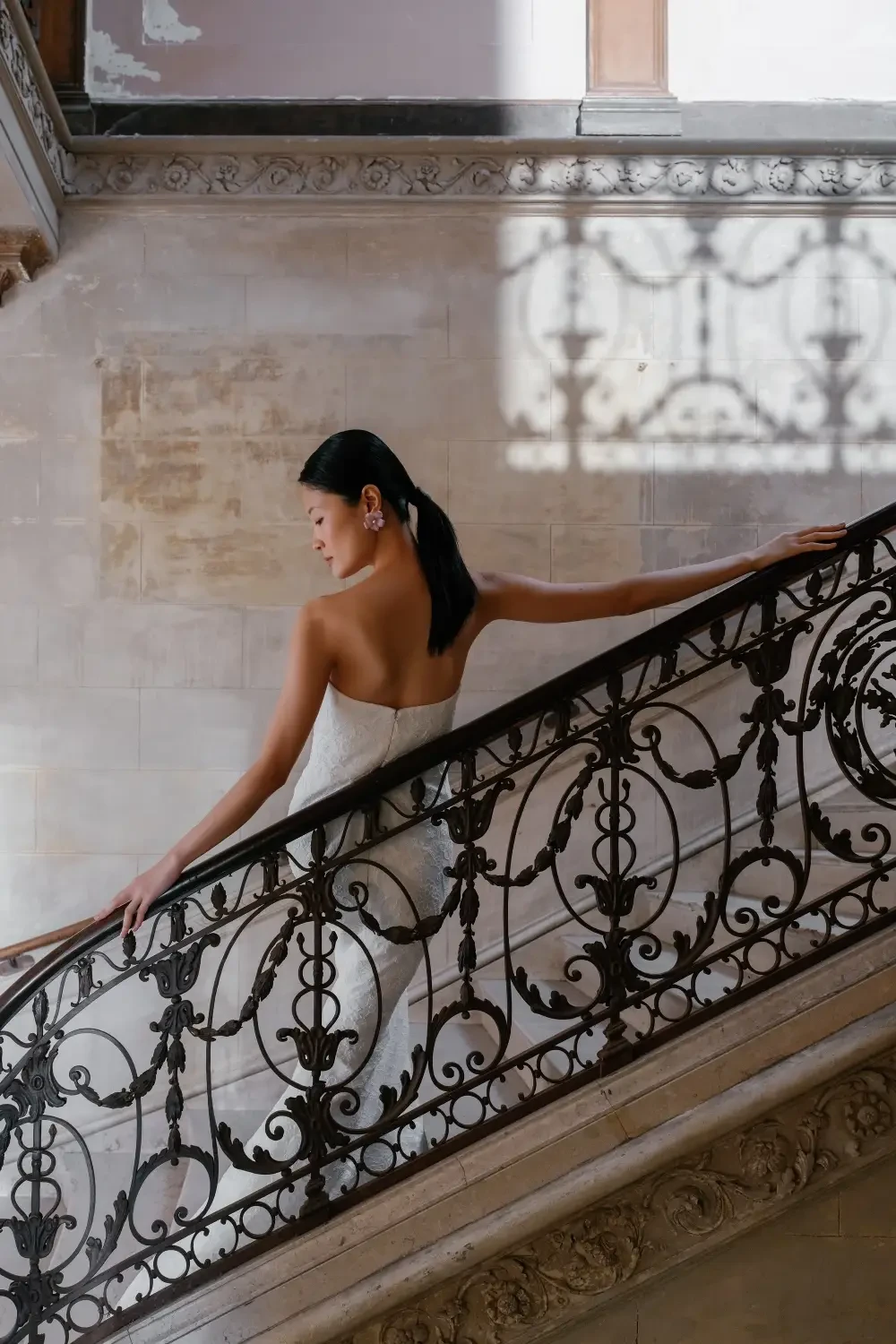 A model wearing a strapless white lace wedding dress stands on a grand staircase inside a historic building.
