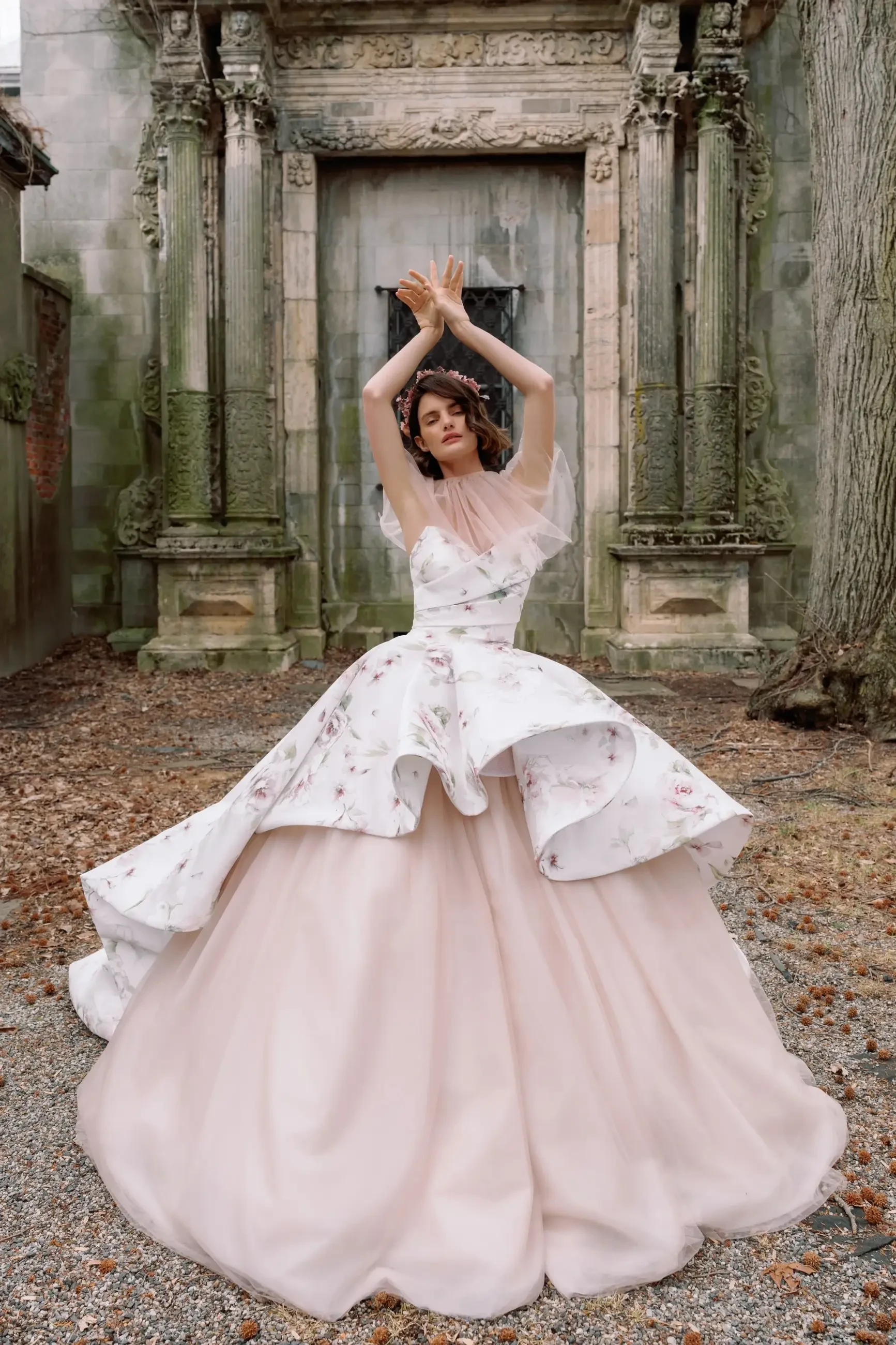 A model poses in a flowing, elegant gown with floral accents, set against a historic stone structure surrounded by gravel and fallen leaves.