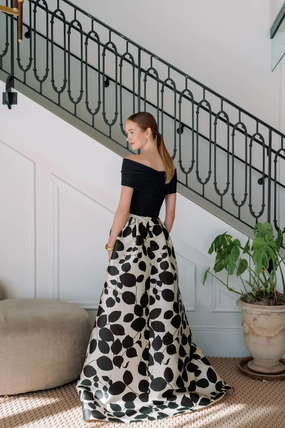 A woman in a strapless black and white polka dot dress stands on a staircase, with a potted plant beside her.