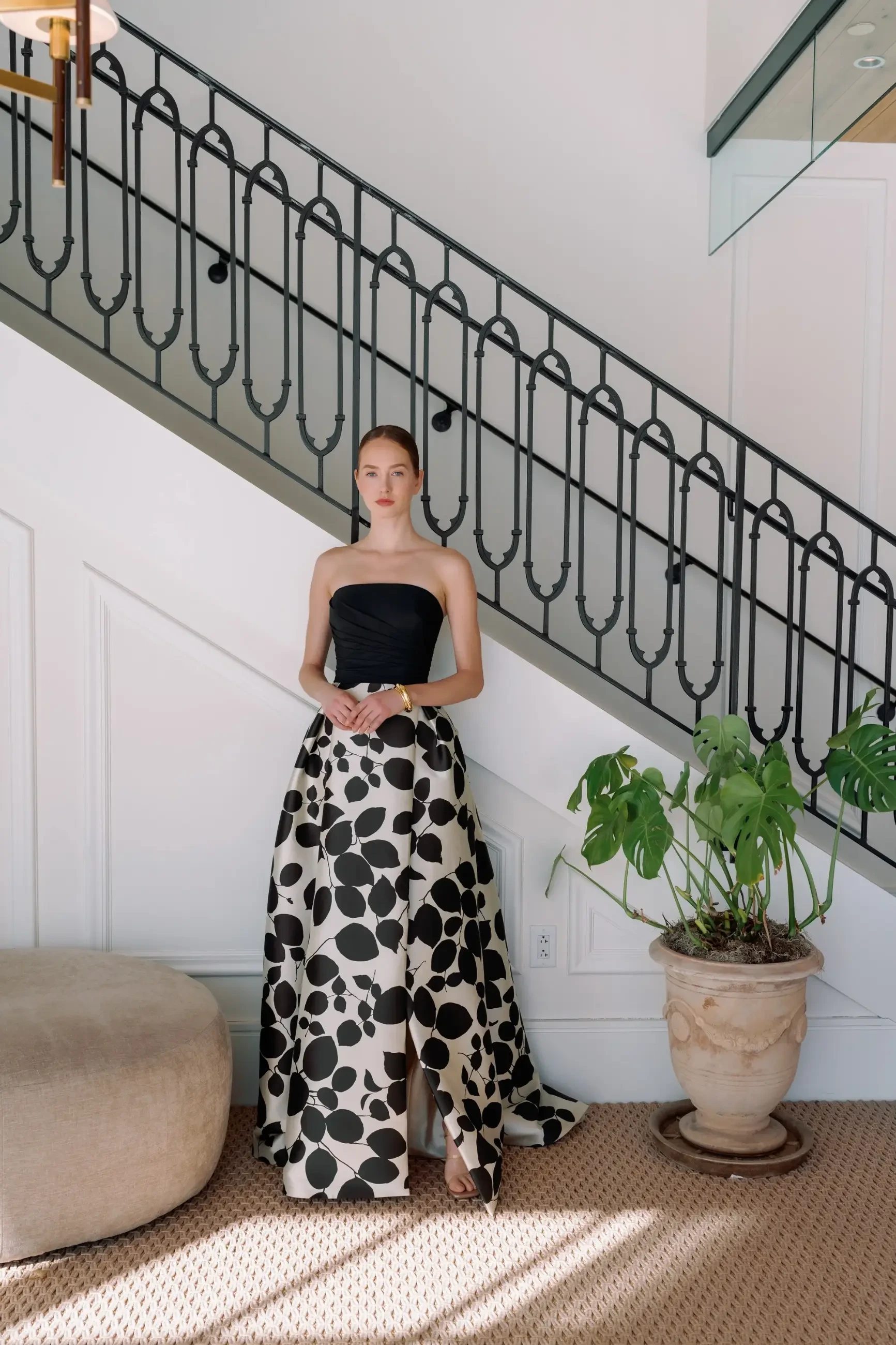 A woman in a strapless black and white dress stands on a staircase, with a decorative plant nearby.