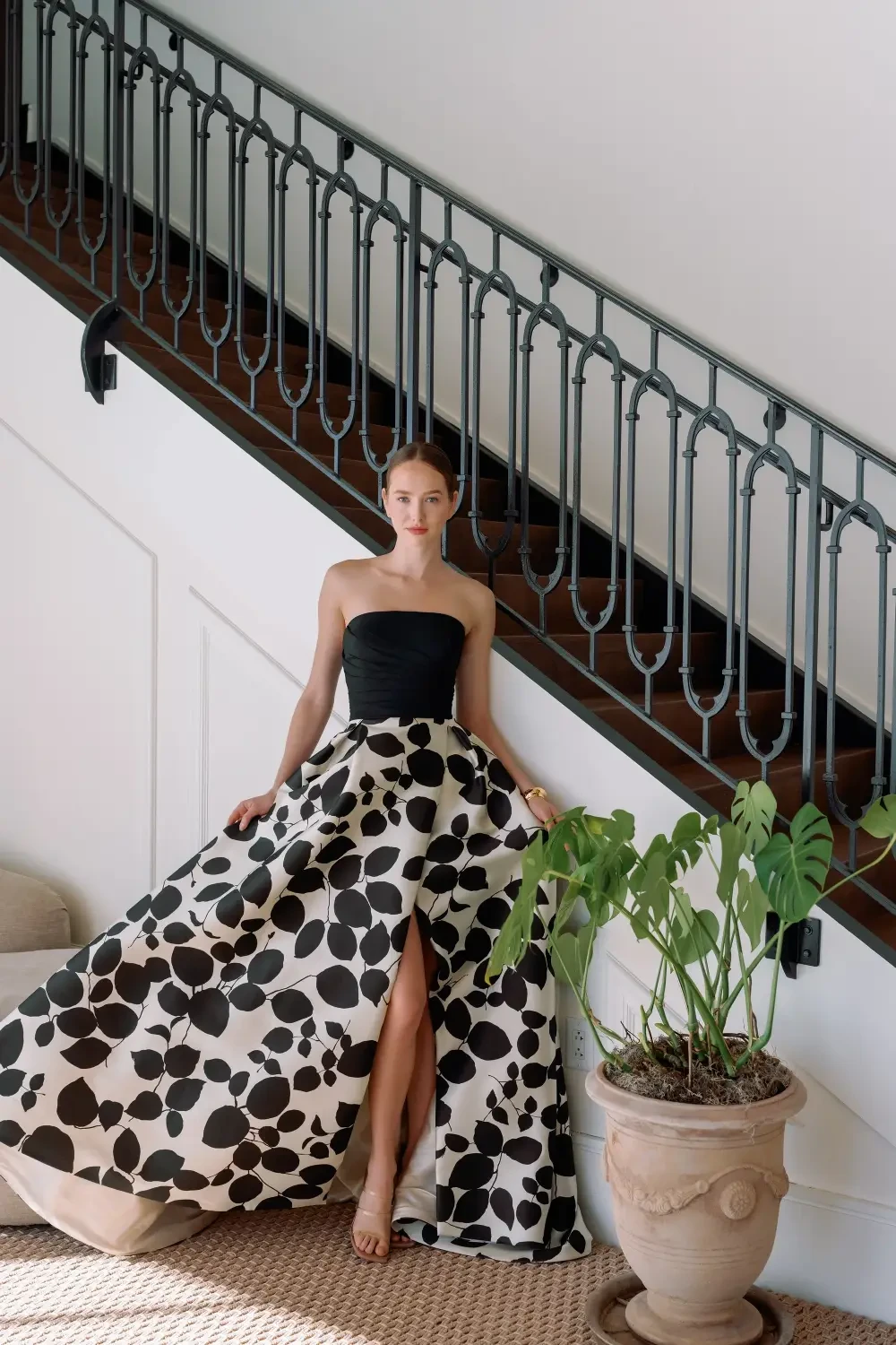 A woman in a strapless black and white polka dot dress stands on a staircase, with a potted plant beside her.