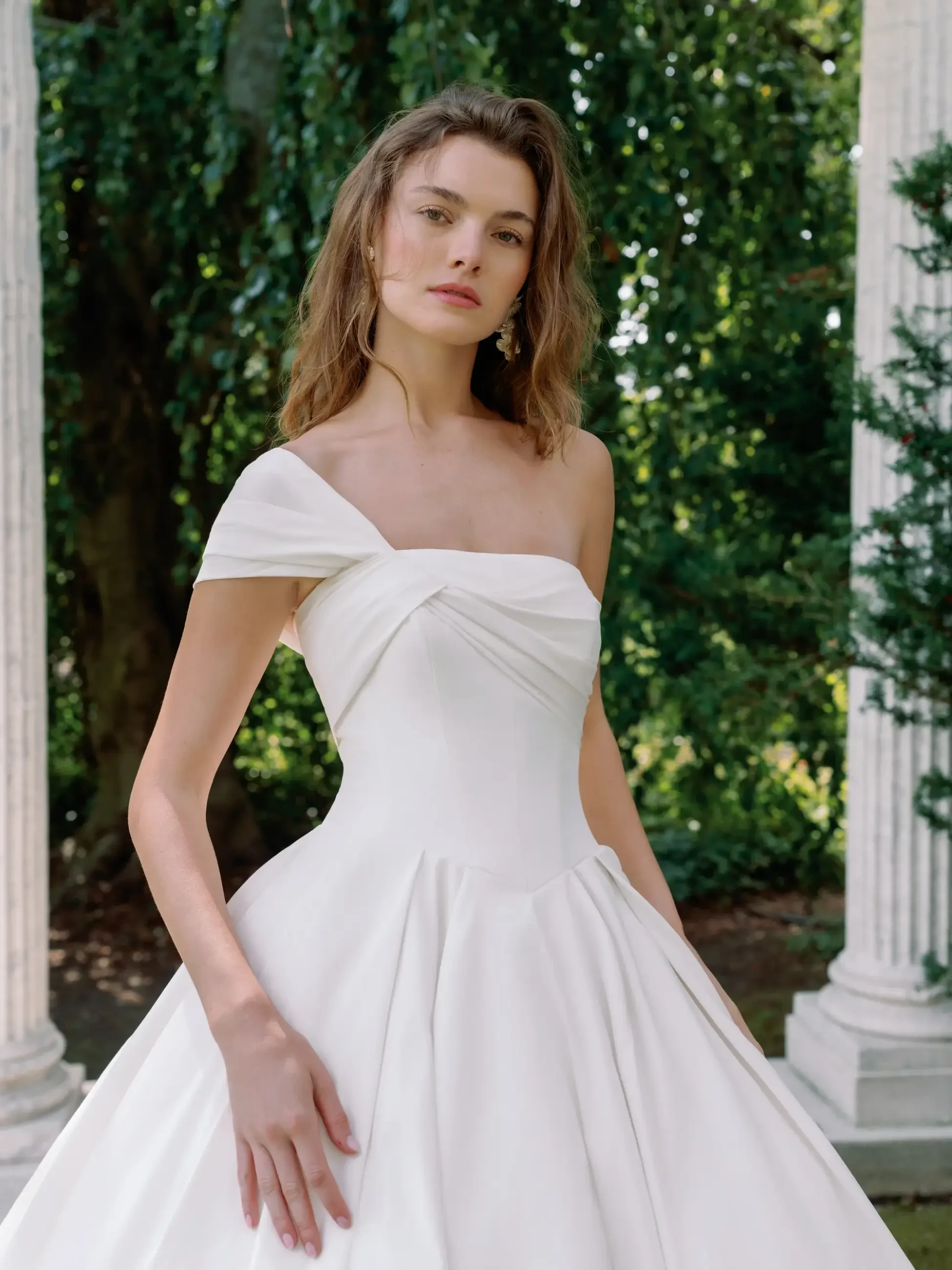 Woman in an elegant, off-shoulder white gown stands outdoors, surrounded by greenery and columns. Her expression is serene and confident.